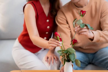 Mutlu Asyalı bir çift vazoda birlikte güller düzenliyor, evde romantik bir sevgililer günü aktivitesi, genç bir adam ve kadın kanepede oturuyor ve birbirlerine bağlanıyorlar, aşk, romantik bir tatil ve ilişki..