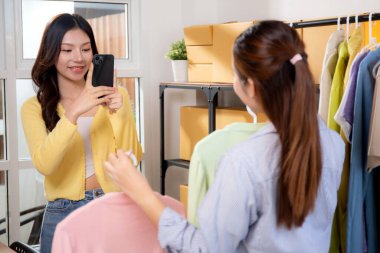İnternetteki mağaza için kıyafet fotoğrafı çeken iki Asyalı kadın, çevrimiçi satıcıyla işbirliği yapan kadın sosyal medyaya fotoğraf giysileri götürüyor, iş kadını veya girişimci ürün satıyor, küçük işletme.