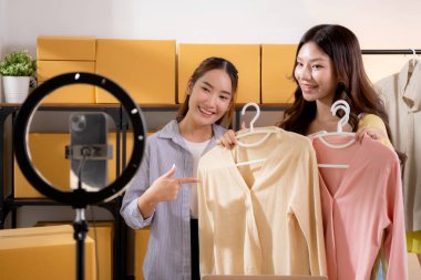 İki genç Asyalı kadın online kıyafet mağazasında canlı yayında canlı yayında sosyal güvenlik telefonuyla kıyafetlerini gösterirken, girişimci online kıyafet satışı ve sunumu için online satıcı,.