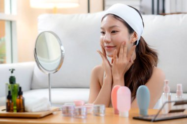 Güzel Asyalı bir kadın, oturma odasında aynaya bakarken kırışıklıklar için yüzüne krem sürüyor. Güzel bir kadın, yanağına nemlendirici sürüyor. Hijyen, cilt bakımı ve güzellik için..