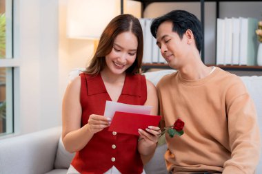 Sevgililer Günü 'nü romantik bir sürprizle birlikte koltukta oturan mutlu Asyalı çift evde gül ve kart okurken, aşkı hissederken, 14 Şubat ve yıldönümlerinde romantik.