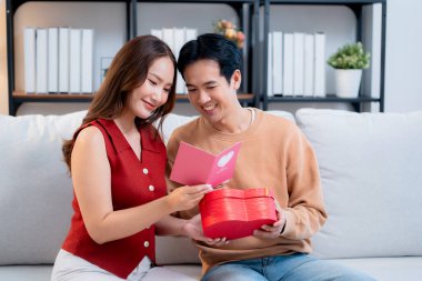 Asyalı genç adam ve kız arkadaş Sevgililer Günü 'nü kutluyorlar. Kartpostal ve hediye kutlamasıyla neşeli bir çift evde kanepede oturup tatil kutluyor ve aşkı hissediyorlar..