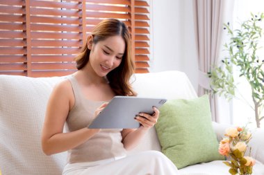 Genç Asyalı iş kadını rahatlamak için tablet bilgisayar kullanıyor ve sosyal medyayı kontrol ediyor. Ev ofisinin oturma odasında kanepede oturuyor. İş kadını tabletle çalışıyor..