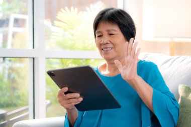 Mutlu üst düzey Asyalı kadın kanepede oturmuş, evdeki tablette, dijital yaşam tarzında, uzaktan iletişimde, yaşlılarla çevrimiçi selamlaşma ya da sosyal medyada rahatlamak için tablet kullanıyor..