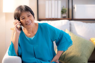 Kıdemli Asyalı kadın telefonda gülümsüyor ve rahatlıyor. Oturma odasındaki koltukta oturuyor. Neşeli, yaşlı bir kadın akıllı telefonuyla evde koltukta oturuyor, yaşam tarzı ve iletişim kuruyor..
