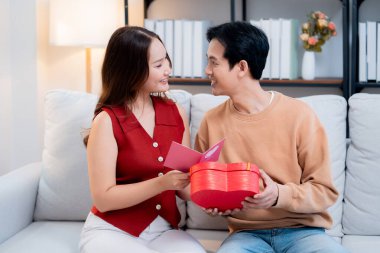 Asyalı genç adam ve kız arkadaş Sevgililer Günü 'nü kutluyorlar. Kartpostal ve hediye kutlamasıyla neşeli bir çift evde kanepede oturup tatil kutluyor ve aşkı hissediyorlar..