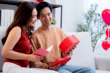 Asyalı genç adam ve kız arkadaş Sevgililer Günü 'nü kutluyorlar. Kartpostal ve hediye kutlamasıyla neşeli bir çift evde kanepede oturup tatil kutluyor ve aşkı hissediyorlar..