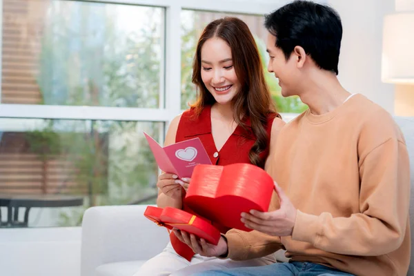Asyalı genç adam ve kız arkadaş Sevgililer Günü 'nü kutluyorlar. Kartpostal ve hediye kutlamasıyla neşeli bir çift evde kanepede oturup tatil kutluyor ve aşkı hissediyorlar..