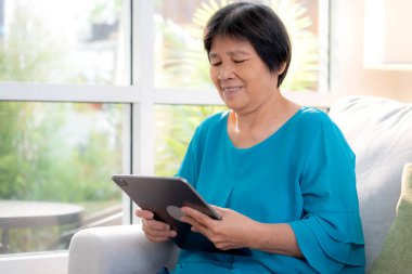 Kıdemli Asyalı kadın evdeki oturma odasında tablet okurken, internet bağlantısı ve dijital yaşam tarzı, sosyal medya için tablet izlerken, uzaktan öğrenirken, sohbet robotu veya yapay zeka kullanırken, kanepede dinleniyor..
