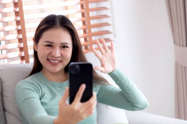 Genç Asyalı bir kadın kanepede oturmuş videolu telefon bekliyor. Oturma odasında telefon görüşmesi yapıyor. Sosyal medyayı internet üzerinden sohbet etmek için kullanıyor..