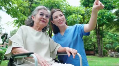 Beyaz kadın bakıcı, bahçede tekerlekli sandalyeli yaşlı hastayı cesaretlendiriyor, profesyonel hemşire, dışarıda rehabilitasyon ve ruh sağlığı terapisi için yaşlı kadına destek oluyor, tıbbi ve yardımda bulunuyor..
