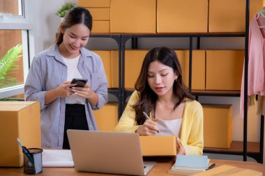 Küçük işletme çevrimiçi mağazasında çalışan iki Asyalı kadın, çevrimiçi satıcı, girişimci iş kadını akıllı telefondan envanter sırasını kontrol et ve merkez ofisteki paketleme için adres yaz.