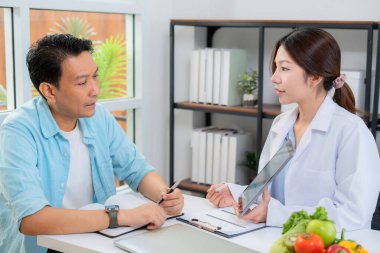 Asyalı beslenme uzmanı, erkek hastaya tablet üzerinde yapay zeka sağlık analizi grafiği, tıbbi veri takibi için teknoloji kullanan doktor, diyet kontrolü, danışmanlık ve beslenme için tıbbi teşhis gösteriyor..