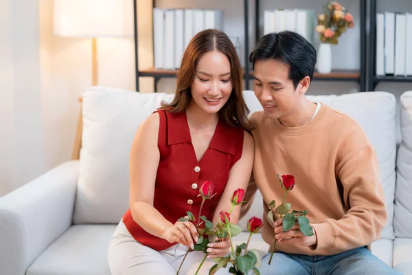 Mutlu Asyalı bir çift vazoda birlikte güller düzenliyor, evde romantik bir sevgililer günü aktivitesi, genç bir adam ve kadın kanepede oturuyor birbirlerine bağlanıyorlar ve aşk, romantik tatil ve ilişki.