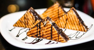 Üstünde çikolata sosu olan dört tabak sandviç. Sandviçler üçgen şeklinde kesiliyor ve beyaz bir tabakta düzenleniyor. Tabak bir masaya konur.