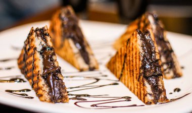 Üstünde çikolata sosu olan üç tabak sandviç. Sandviçler üçgen şeklinde kesiliyor ve beyaz bir tabakta düzenleniyor. Tabak bir masaya konur.