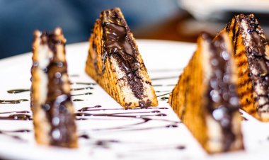 Üstünde çikolata sosu olan üç tabak sandviç. Sandviçler üçgen şeklinde kesiliyor ve beyaz bir tabakta düzenleniyor. Tabak bir masaya konur.