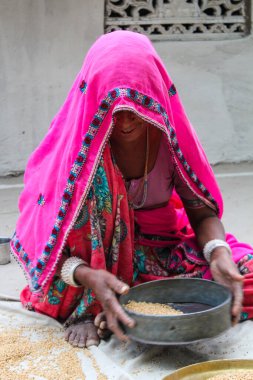 Pembe sari giyen bir kadın yerde oturuyor ve elinde bir kase tutuyor. Kolye ve bilezik takıyor.