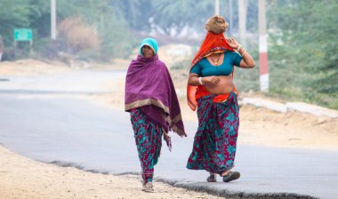Renkli giysiler içinde yolda yürüyen iki kadın. Kadınlardan biri mavi bir bluz giyiyor.