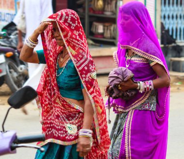 Mor ve kırmızı giysili iki kadın caddede yürüyor. Kadınlardan biri bir çanta tutuyor.