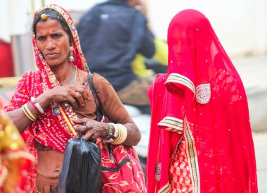 Kırmızı sari giyen iki kadın. Bir kadın siyah bir çanta tutuyor. Diğer kadın bir çanta tutuyor.
