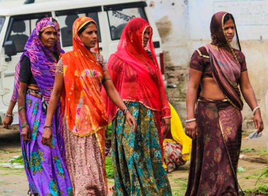 Renkli sariler giyen dört kadın caddede yürüyor. Kadınlardan biri kırmızı eşarp takıyor.
