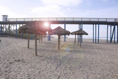 beach vacation background with palm tree umbrellas and pier in summer with sun flare in rosarito mexico clipart