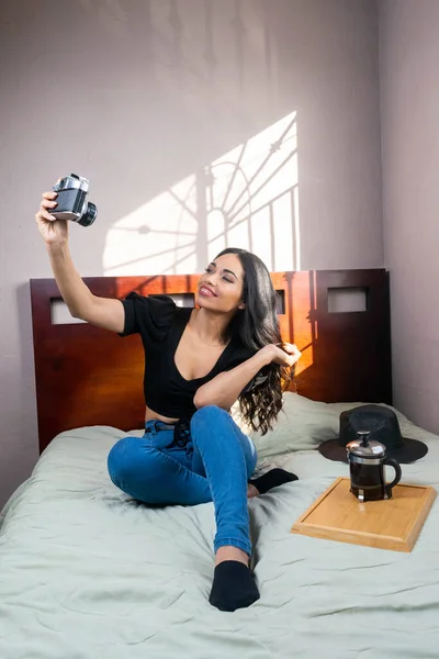 Relaxed young woman in bed with coffee after work taking pictures with old camera