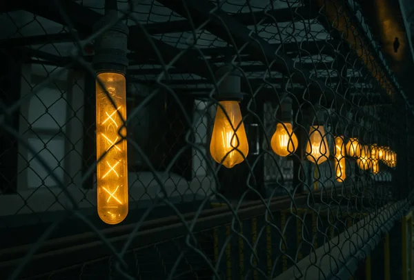 Farklı uzun ve yuvarlak şekilli lambalar ızgaranın arkasında sarı ışık ile parlıyor. İplik lambada görülebilir. Gergin atmosfer.