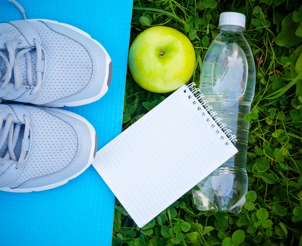 Scarpe sportive sneakers su tappetino yoga, bottiglia d'acqua e mela su erba fresca verde Foto Stock