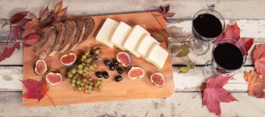 İki kadeh kırmızı şarap, incir, peynir, üzüm, ekmek, siyah zeytin ve yıllanmış ahşap masada sonbahar yaprakları. Üst görünüm.