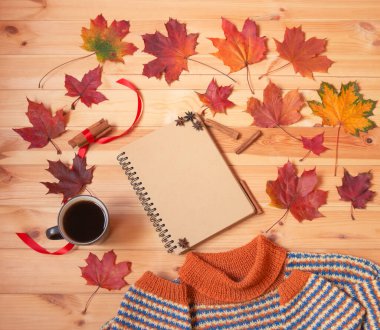 Boş defter, sıcak kahve kupası, renkli sonbahar akçaağaç yaprakları ve ahşap yüzeyde ev yapımı sıcak süveter. Üst görünüm, alanı kopyala.