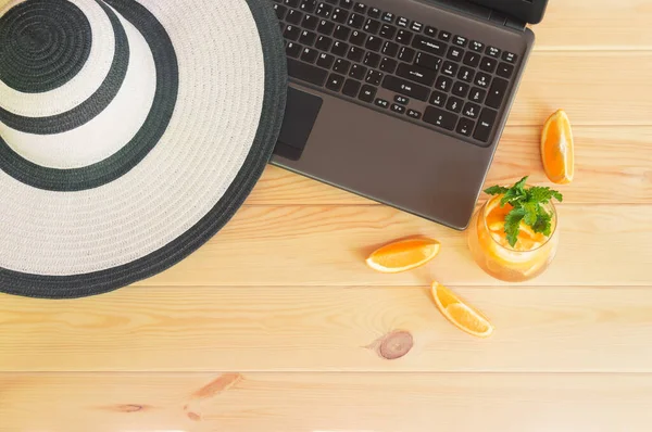 Summer hat, laptop and orange lemonade on wooden table. Summer holidays concept. Top view.