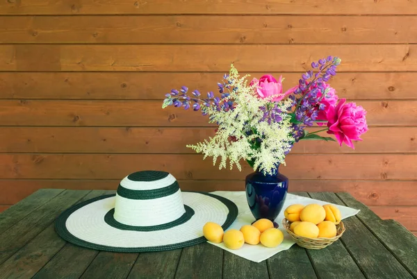Beautiful bouquet made of white astilbe, lupine and pink peonies in blue vase, summer hat and fresh ripe apricots in basket and near on rustic wooden table