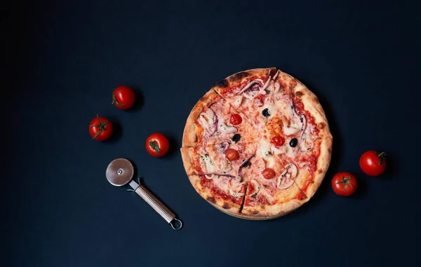 Delicious seafood pizza, pizza cutter and fresh tomatoes on dark background. Top view, copy space.