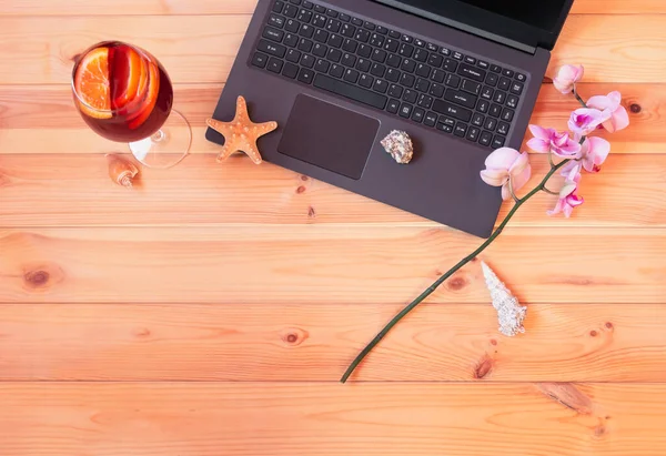 Laptop, glass of sangria, branch of orchid, starfish and seashells on wooden table. Summer holidays or freelance concept. Top view, copy space.