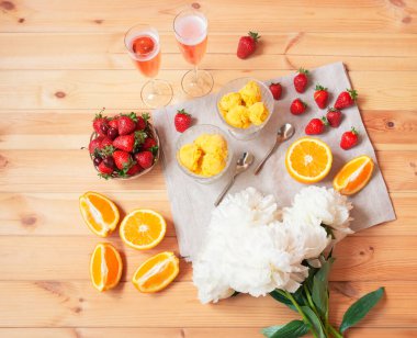İki bardak gül şampanyası, portakallı dondurma, taze portakallar, beyaz şakayıklar ve ahşap masada taze çilek. Üst görünüm.