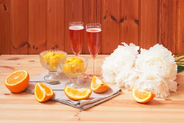 Glasses of rose champagne, white peony flowers and bowls of orange sorbet ice cream on wooden table. Selective focus. View with copy space.