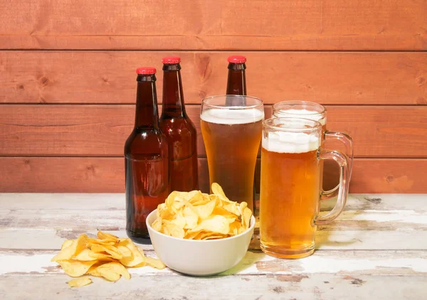 Bir bardak bira, bir kupa bira, bir şişe bira ve yıllanmış ahşap masada patates cipsi ve kase..