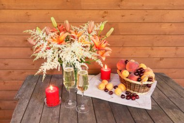 Zambaklar ve cam vazoda astilbe çiçekleri, iki bardak şampanya, kırmızı mumlar ve olgun meyvelerle dolu sepet. Seçici odak.