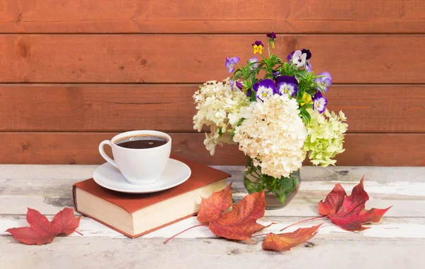 Cup of coffee, book, autumnal white hydrangea and pansy flowers in glass vase and autumn maple leaves  on aged wooden table. View with copy space.