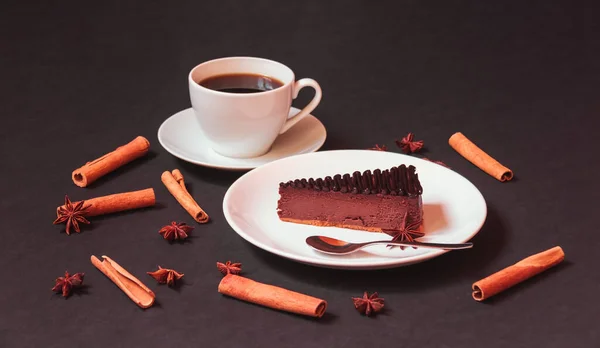 Chocolate cheesecake with chocolate sauce, spices and cup of coffee on dark background. Selective focus on cheesecake.