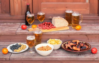 Bira bardakları ve kupalar, bira şişesi, bira aperatifleri, ızgara tavuk kanatları, ekmek ve küçük sucuklu sosisler kırsal ahşap merdiven basamaklarında..