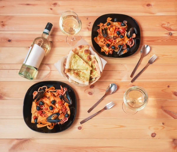 Plates of pappardelle pasta with seafood with tomato sauce, bottle of white wine, glasses of white wine and focaccia bread on wooden table. Top view.