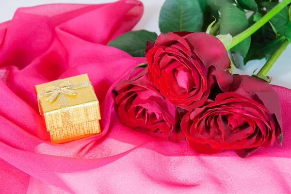 Goldene Geschenkschachtel und drei rote Rosen auf einem rosa Seidenschal — Stockfoto
