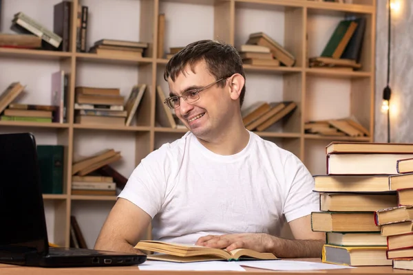 young male student sits at a table with books and smiles into a laptop on the background of a cabinet. European appearance. distance learning. preparation for exams.