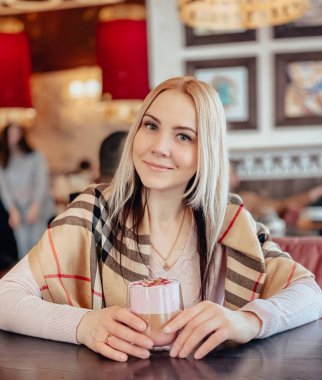 Omzunda yumuşak pembe bir kazakla sarışın bir kız rahat bir restoranda oturur ve rahatlar ve kahve içer ve masasında bir parça kek yer..