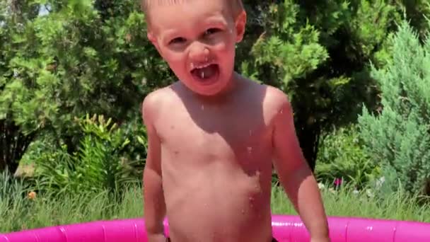Маленький мальчик в надувном бассейне для детей, веселый, улыбающийся, брызгает водой — стоковое видео