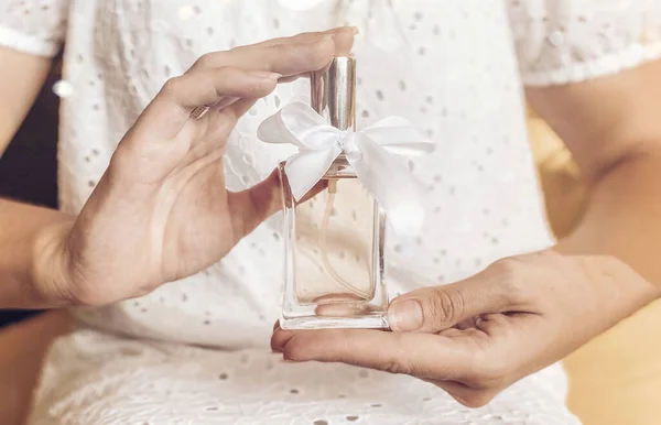 Bottiglia di profumo con un nastro regalo bianco in una mano femminile sullo sfondo di una ragazza in un abito bianco in colori chiari pastello. — Foto Stock