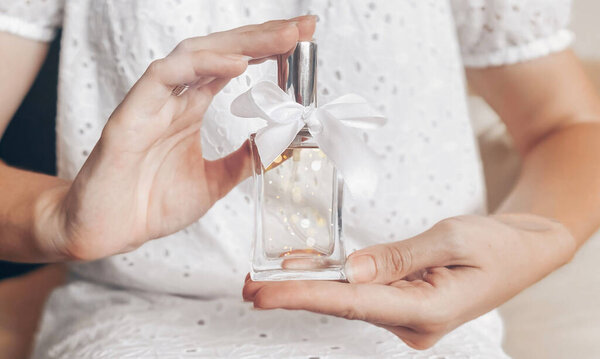 Бутылка духов с белой лентой подарка в женской руке на фоне девушки в белом платье в пастельных светлых тонах.